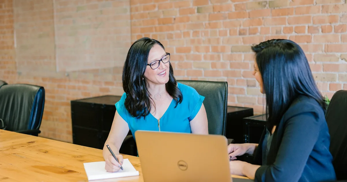 Two women in tailored workwear talking across a conference table