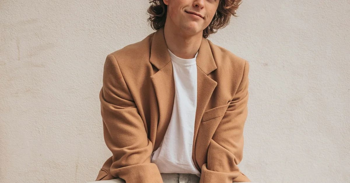 Man in a camel overcoat over a white t-shirt sitting against a wall — the one-notch lift this report measures